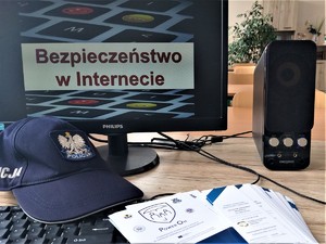 Klasa szkolna. Biurko a na nim komputer z wyświetlonym na ekranie slajdem z napisem Bezpieczeństwo w Internecie. Obok głośnik, czapka policyjna, ulotki.