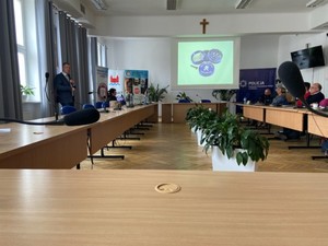 Sala w urzędzie. Stoły ustawione w &amp;quot;podkowę&amp;quot;. Przy nich siedzą osoby. W tle mężczyzna przemawiający, w tle rollup między innymi  KPP w Nidzicy, sanepidu i starostwa nidzickiego. Na ścianie wyświetlony slajd prezentacji.