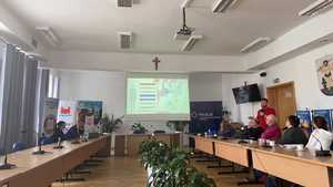 Sala w urzędzie. Stoły ustawione w &amp;quot;podkowę&amp;quot;. Przy nich siedzą osoby. W tle mężczyzna przemawiający, w tle rollup między innymi  KPP w Nidzicy, sanepidu i starostwa nidzickiego. Na ścianie wyświetlony slajd prezentacji.