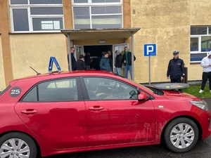 Na pierwszym planie czerwone auto osobowe. Za nim kilka osób, m.in. policjant ruchu drogowego. W tle budynek z żółtą elewacją.