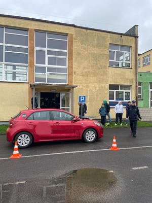 Na pierwszym planie czerwone auto osobowe. Za nim kilka osób, m.in. policjant ruchu drogowego. W tle budynek z żółtą elewacją.