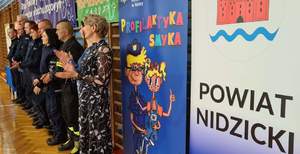 Zdjęcie grupowe policjantów i przedstawicieli władz samorządowych oraz roll up &amp;quot;Profilaktyka smyka&amp;quot; i powiat nidzicki.