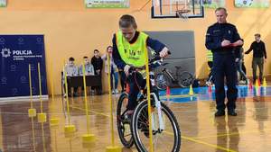 Sala sportowa. Zawodnik w żółtej kamizelce pokonujący jedną z przeszkód na torze. W tle policjant.
