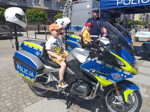 2 dzieci siedzących na motocyklach policyjnych. Obok stoi policjant.