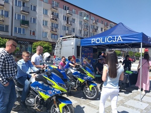 Plac w mieście. Na pierwszym planie stoją 2 policyjne motocykle, za nimi namiot policyjny a pod nim stolik z upominkami, wokół ludzie .