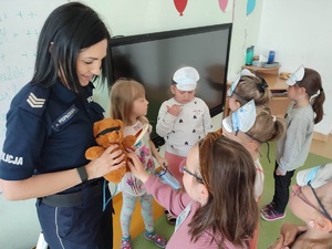 Sala lekcyjna. Stojąca policjantka, która trzyma w ręku misia. Wokół niej stoją dzieci.