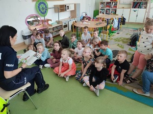Sala lekcyjna. Na krzesełku siedzi policjantka i czyta książkę dzieciom siedzącym przed nią na dywanie.