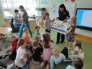 Sala zajęciowa. Policjantka stoi. obok niej stoi 2 dzieci trzymających plakat z rowerem. przed nimi znajduje się grupa dzieci.