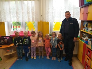 Policjant z grupą przedszkolaków stoją pod ścianą w sali zajęciowej.