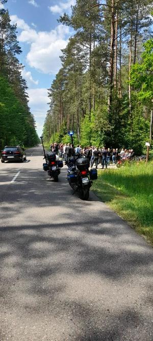 Pora dzienna droga przez las. Na niej stoją policyjne motocykle. Na poboczu stoją osoby.
