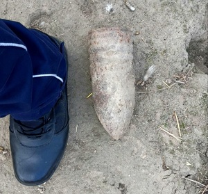 Skorodowany pocisk artyleryjski. Po prawej stronie noga w czarnym bucie. Wszystko leży na piaszczystym podłożu.