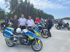 Policjanci ruchu drogowego stojący przy motocyklach. W tle motocykle i motocykliści oraz las i duży betonowy plac.