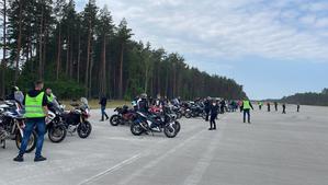 Motocykliści i motocykle stojące na dużym wybetonowanym placu. W tle las.