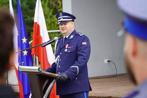Komendant Powiatowy Policji w Nidzicy w trakcie przemowy. W tle 2 flagi Polski i flaga UE.