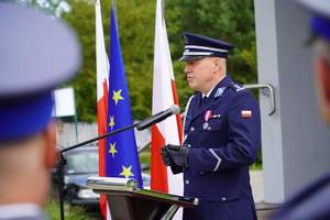 Zastępca Komendanta Wojewódzkiego Policji w Olsztynie  w trakcie przemówienia. Stoi za mównicą, za nim 2 flagi Polski i flaga UE.