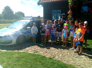 Zdjęcie grupowe. Policjant, policjantka kilkanaścioro dzieci. Po lewej stronie stoi radiowóz. W tle drewniany ciemny budenek, na nim żywe kwiaty i niebieskie okno.