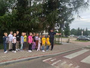 Grupa kilkanaściorga dzieci z policjantem. Wszyscy stoją przed przejściem dla pieszych policjant wskazuje na znak informacyjny dot. przejścia dla pieszych.