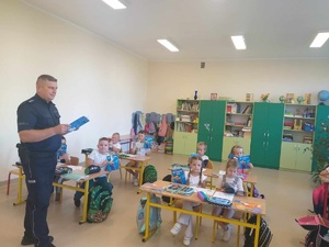 Klasa. Dzieci siedzące w ławkach. Po lewej stronie stoi policjant trzymający książkę w rękach.