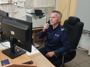 Policjant siedzący za biurkiem, na którym stoi monitor i leżą dokumenty. Policjant trzyma w prawym ręku czarną słuchawkę telefonu.
