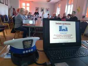 Na pierwszym planie monitor komputera z wyświetlonym slajdem dot. debaty, obok leży czapka policjanta ruchu drogowego. W tle siedzący przy stole uczestniczy debaty.