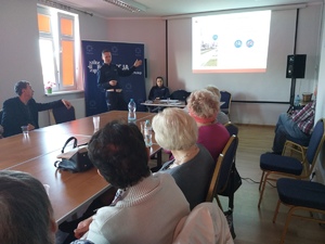 Seniorzy siedzący przy stole. W tle policjant i slajd prezentacji nt. ruchu drogowego.