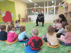 Przedszkole. dzieci siedzą na dywanie. Przed nimi na małym krześle siedzi policjant czytający bajkę.