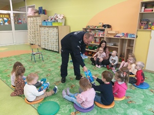 Przedszkole. Policjant rozdaje książki siedzącym na dywanie  przedszkolakom.