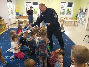 Policjant rozdaje książki siedzącym i stojącym na dywanie przedszkolakom.