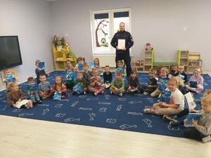 Zdjęcie grupowe. Przedszkolaki siedzą na dywanie a za nimi stoi policjant trzymający w rękach podziękowanie.