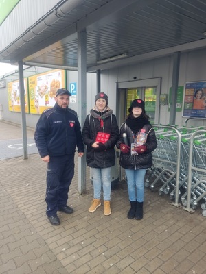 Policjant i 2 wolontariuszy WOŚP, w tle wejście do sklepu.