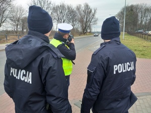 Policjant ruchu drogowego w trakcie mierzenia prędkości jazdy. Za nim stoi 2 policjantów kursu podstawowego.