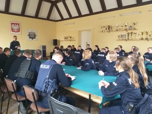 Słuchacze Akademii Policji w Szczytnie i policjanci KPP w Nidzicy siedzą na świetlicy przy stołach przykrytych zielonym obrusem. W tle na żółtej ścianie półki z pucharami. Trwa odprawa do służby.