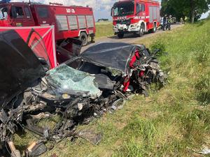 Miejsce wypadku drogowego ze skutkiem śmiertelnym. Auto rozbite na skutek zderzenia znajdujące się na trawie poza droga. Na drodze stoją wozy strażackie i czerwony parawan.
