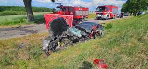 Miejsce wypadku drogowego ze skutkiem śmiertelnym. Auto rozbite na skutek zderzenia znajdujące się na trawie poza droga. Na drodze elementy pojazdów. W tle wozy straży pożarnej, policja.
