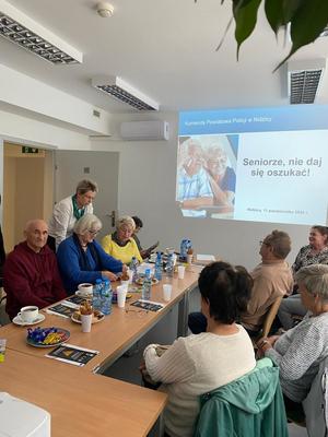 &quot;Dzień Seniora&quot; - spotkanie z seniorami. Na ścianie wyświetlony slajd. Seniorzy siedzą przy stole.