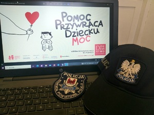 Spotkania nidzickich policjantów na temat przemocy podczas „Tygodnia świadomości przemocy domowej na Warmii i Mazurach”. Ekran laptopa z wyświetlonym plakatem. Na klawiaturze leży czapka policyjna i naszywka KPP w Nidzicy.