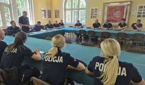 Policjanci podczas odprawy służby siedzą na dużej sali przy stołach przykrytych zielonym materiałem.