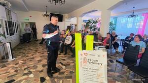 seniorzy siedzący na krzesłach i przemawiający do nich policjant trzymający odblask